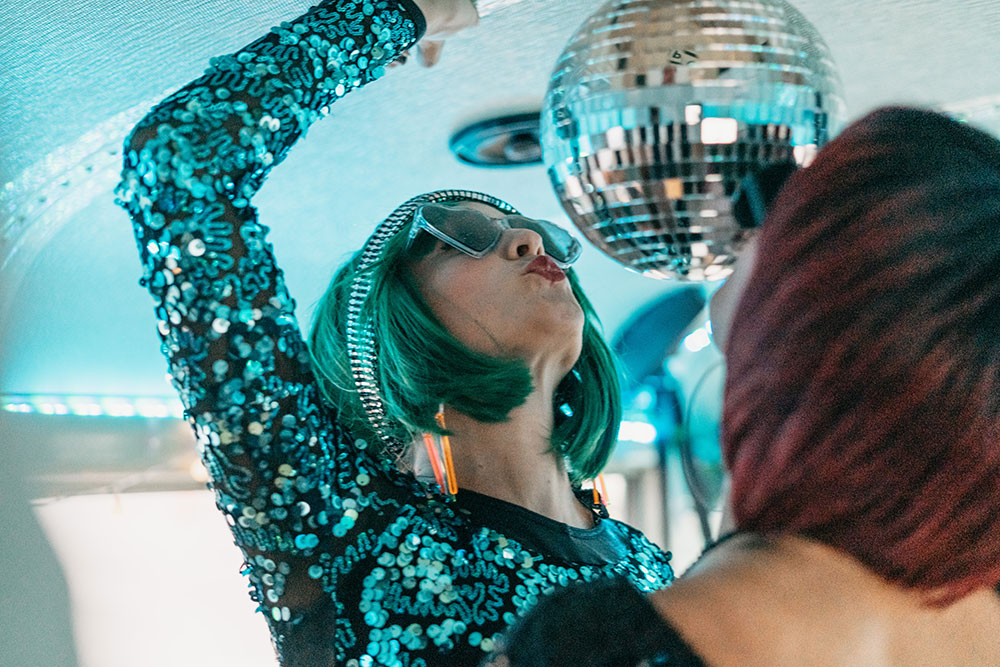 Guest in green hair and sequins reaching for a disco ball on the party bus