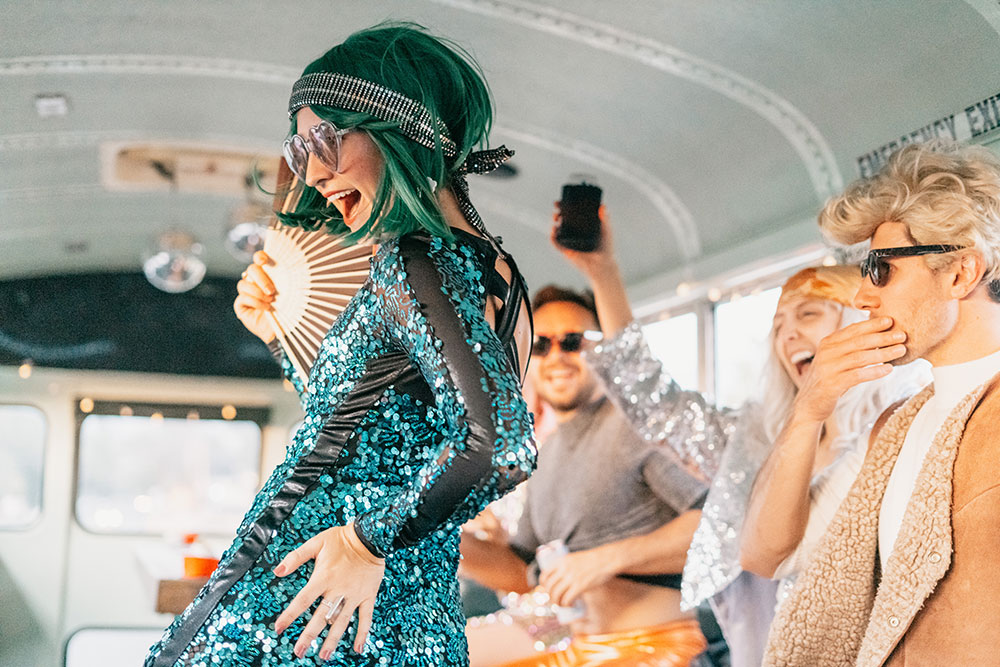 Guest in a green wig and sequin dress dancing with friends on the bus