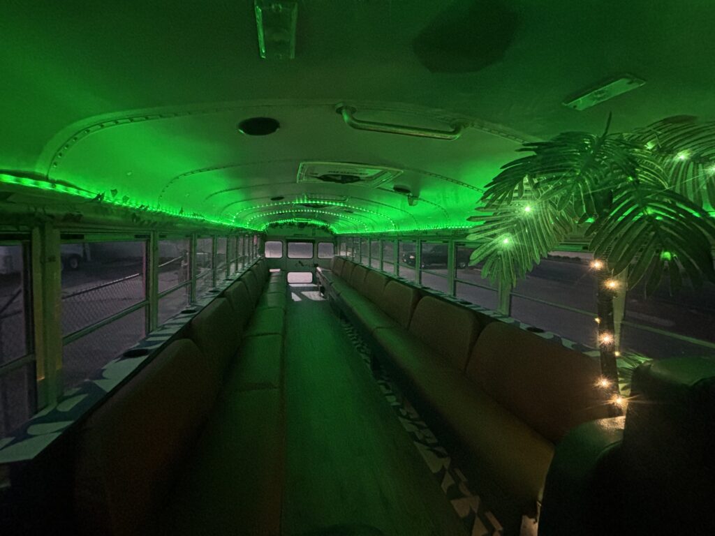 The interior of a bus, designed for party events with green lighting and a imitation palm tree adorned with christmas lights.